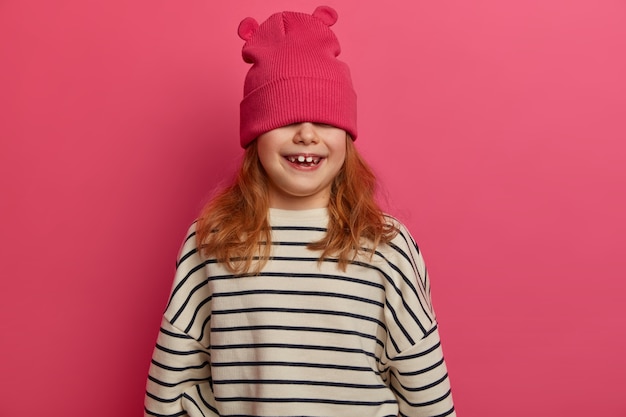 Free photo carefree adorable girl plays indoor, enjoys great day, expresses positive attitude, hides face with hat, wears loose jumper with black stripes, poses against pink wall. children, fun concept