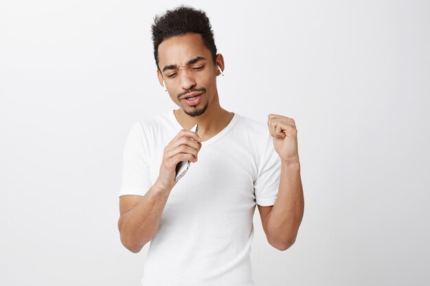 Carefree african-american guy playing karaoke, singing in smartphone mic, wearing wireless headphones