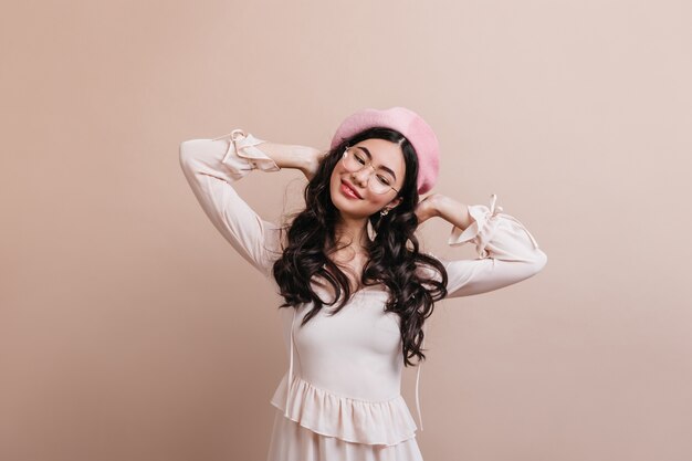 Carefree asian woman wearing french beret. Positive long-haired chinese woman standing on beige background.