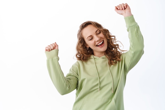 Carefree attractive girl dancing, having fun and enjoying cool music, raising hands up to beat of song, posing upbeat against white wall