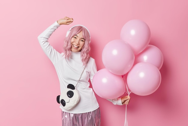 Free photo carefree birthday girl dances with rhythm of music listens audio track via headphones holds bunch of inflated balloons isolated over pink background enjoys party time. people and celebration