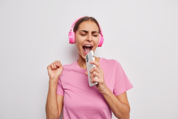 Carefree cheerful woman sings song holds smartphone near mouth as if microphone has fun foolishes around dressed in casual pink t shirt isolated over white background Music and entertainment