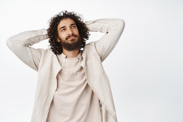 Free photo carefree curlyhaired man holding hands behind back relaxing and smiling lying and resting standing over white background