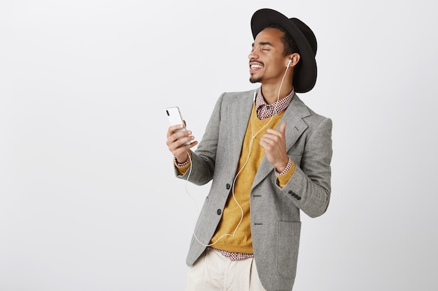 Carefree dancing african-american man listening music in headphones, smiling and holding smartphone