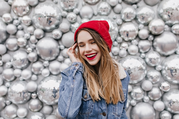 Carefree female model with bright makeup posing with cute smile on shiny wall. Charming caucasian woman in red hat and denim jacket smiling near disco balls.