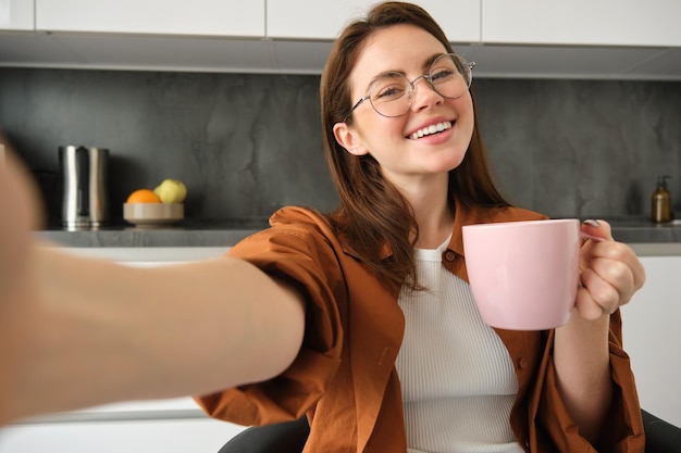 Free Photo carefree girl in glasses takes selfie with smartphone smiling and looking happy enjoying hot drink