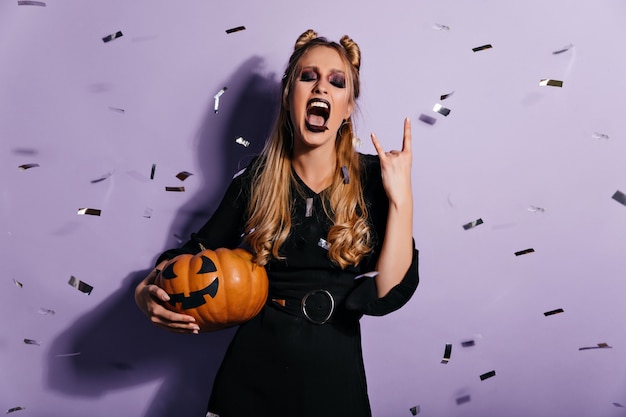 Carefree girl in halloween costume preparing for party. Cute witch with pumpkin standing under confetti.