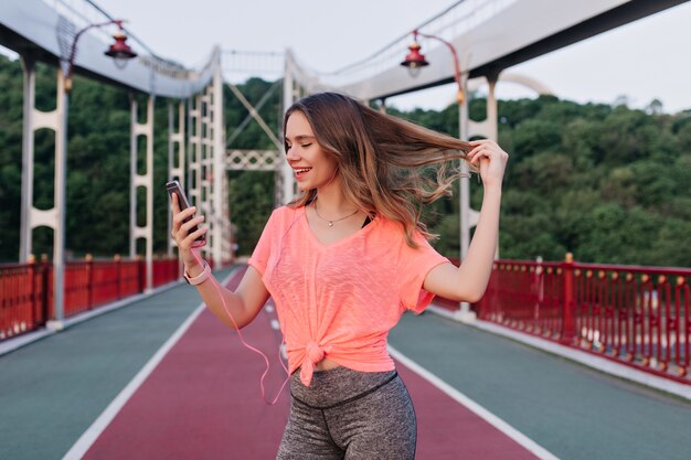 Carefree girl playing with her hair while taking picture of herself at stadium. Amazing caucasian woman making selfie with smartphone.