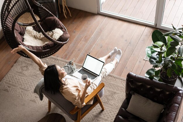 Carefree happy asian girl finish working and looking satisfied sitting at home with laptop computer ...