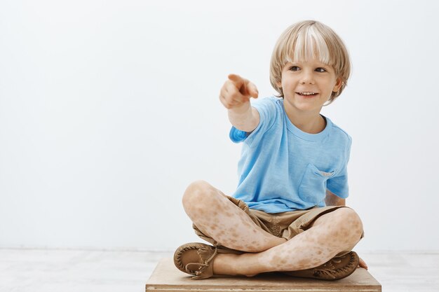 carefree happy blond child being entertained, sitting with crossed feet and pointing aside while smiling broadly, being interested and curious