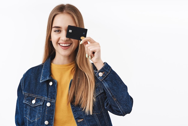 Carefree and happy smiling girl showing credit card near eye finally receive payment gonna waste money place cash on deposit using cashback making investment stand white background