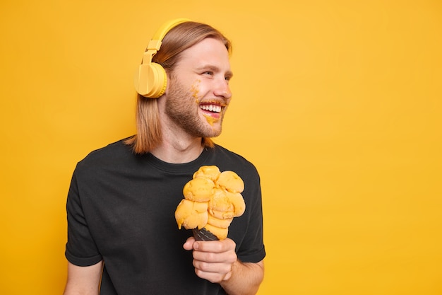 Carefree hipster guy with ginger hair smiles positively focused away has fun eats tasty ice cream has dirty face listens music via headphones poses against yellow wall copy space on right