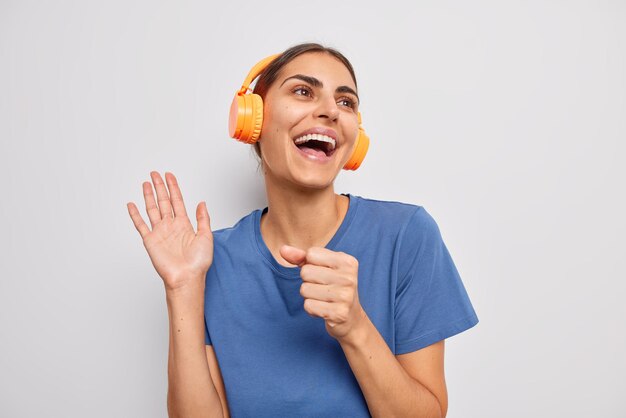 Carefree optimistic woman keeps palm raised hand near mouth as if microphone sings favorite song along dressed in casual blue t shirt isolated over white background uses wireless orange headphones