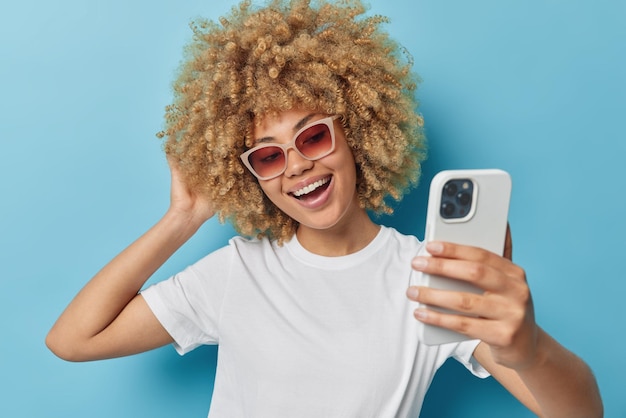 Free Photo carefree optimistic young woman has fair curly hair has cheerful expression takes selfie via smartphone wears sunglasses and casual white t shirt isolated over blue background makes video call