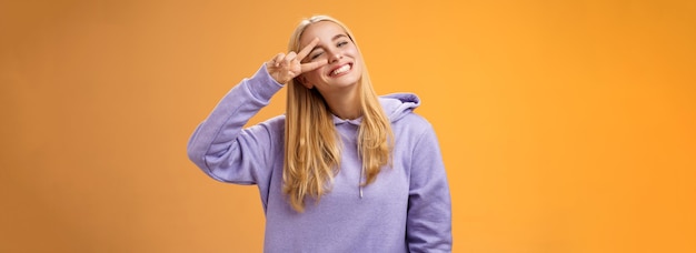 Free photo carefree optimsitic young cute blond caucasian girlfriend having fun enjoying sun perfect spring weather winking cheeky smiling tilting head happily show peace victory sign orange background