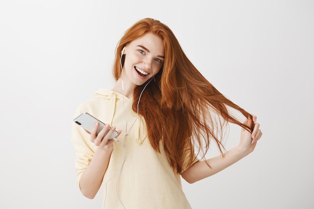 Carefree redhead girl listening music in earphones and using smartphone