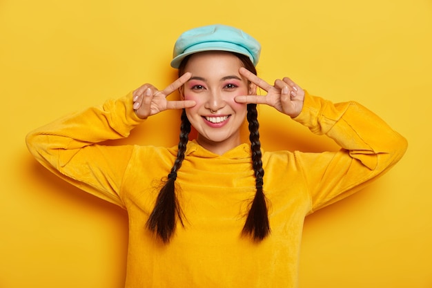 Free photo carefree smiling asian lady makes victory peace gesture near eyes, has happy mood, smiles gently, wears vivid makeup, wears stylish hat and sweatshirt, isolated on yellow wall