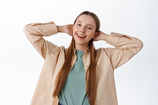 Carefree teenage female student holding hands behind head, smiling while relaxing, resting, having leisure time, enjoying weekend free time, standing over white wall