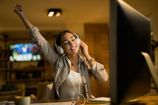 Carefree woman with eyes closed enjoying in music over headphones while using computer in the evening at home