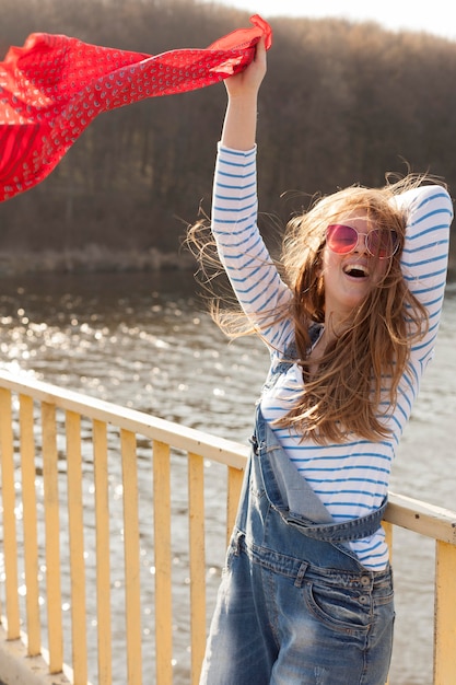 Free Photo carefree woman with sunglasses holding scarf in the wind by the lake