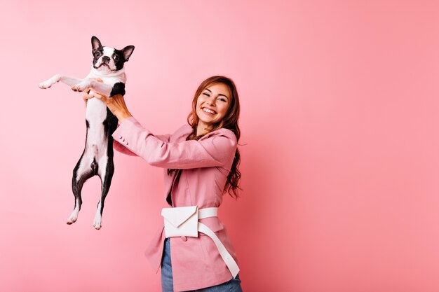 Carefree young lady holding black french bulldog with sincere smile. Indoor portrait of joyful girl playing with dog on pastel.