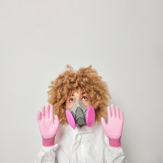 Free Photo careful woman keeps palms towards camera to protect herself got in ecological trouble doesnt have any fresh air to breath wears white proective suit gas mask and gloves poses indoor looks above