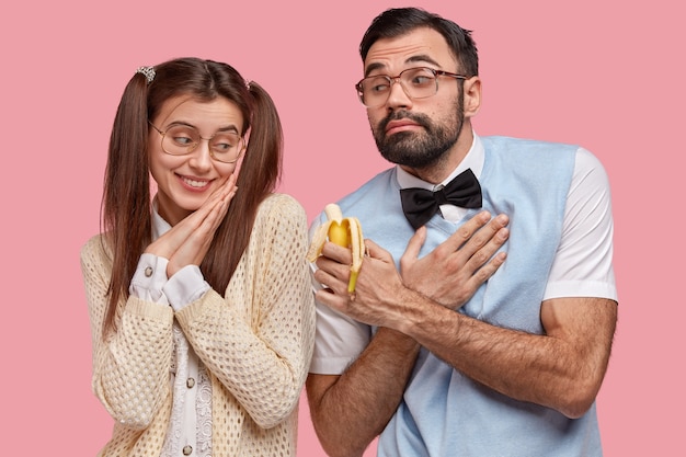 Caring boyfriend with thick stubble, dressed elegantly, looks awkward, keeps hand on chest, suggests girlfriend to bite banana 