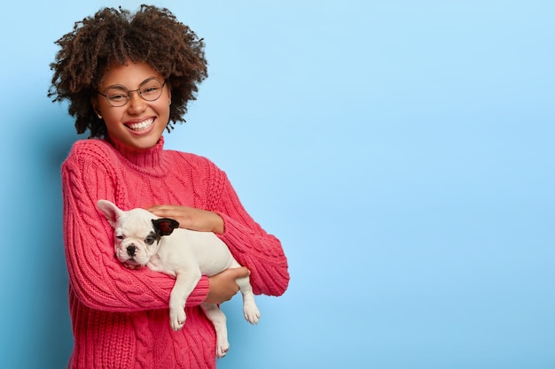 Free Photo caring dark skinned animal owner holds small puppy, likes pets, wears spectacles and pink sweater, smiles gladfully