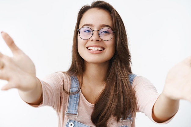 Caring tender and attractive feminine woman in glasses extending arms towards camera to give kiss and hug wanting hold in arms thing smiling broadly standing delighted and cute over gray wall