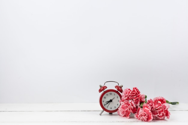 Free Photo carnation red flowers and red alarm clock on white wooden surface