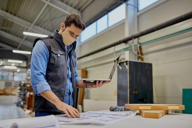 Carpenter with face mask examining blueprints while using computer at woodworking production facility