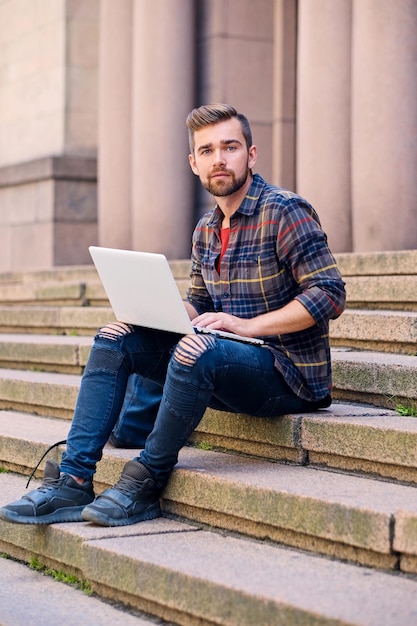 A casual bearded male dressed in a jeans and fleece shirt sits on a step and using a laptop.