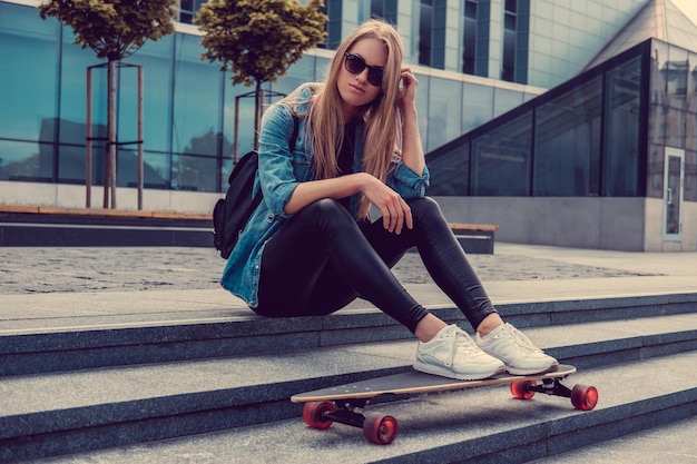 Casual blond female in denim shirt posing on stairs with longboard over glass building.