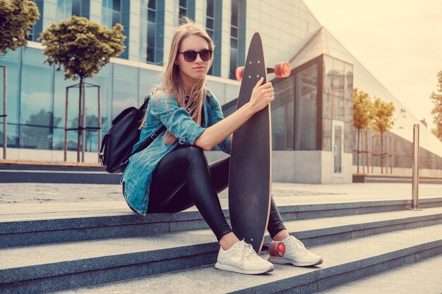 Casual blond female in denim shirt posing on stairs with longboard over glass building.