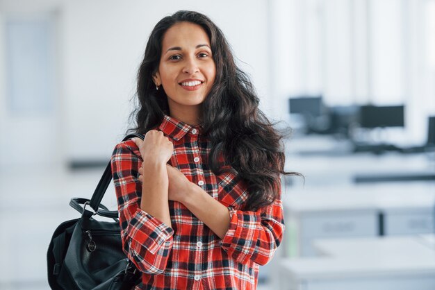 In casual clothes. Portrait of attractive young woman standing in the office with black bag