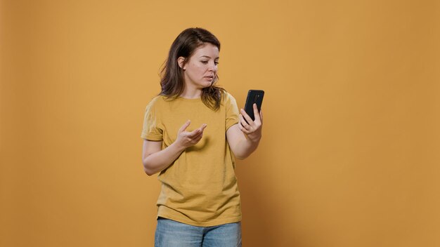 Casual happy woman in video call conference using smartphone waving hello at front camera in studio. Smiling person fixing hair and having online conversation using mobile phone messaging app.