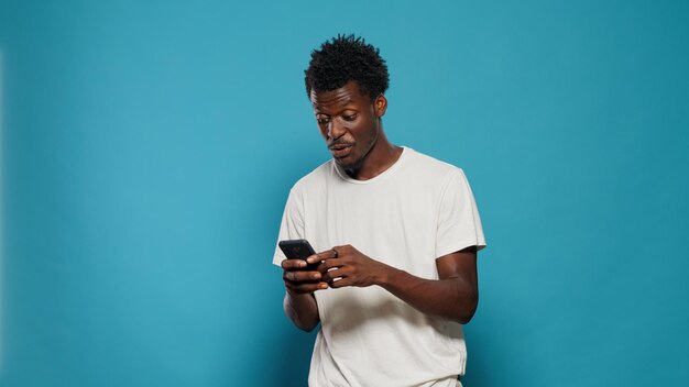 Casual person looking at smartphone and dancing in studio. Joyful man listening to music and song while holding phone with touchscreen in hand, standing over isolated background.