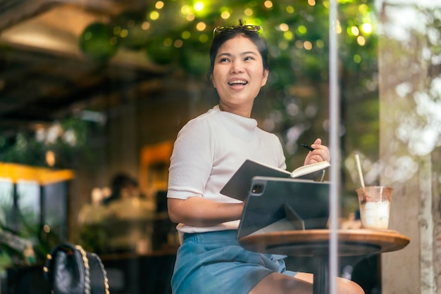 Casual relax asian female freelance digital nomad hand writing sketch book ideas noted for her new project at cafe shot through coffeeshop window glassasian female sit work in cafe cheerful feeling