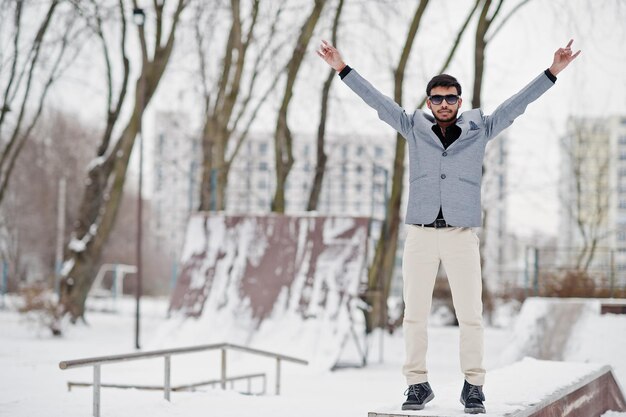 Casual young indian man in silver jacket and sunglasses showing rock finger sign hands up and posed at winter day