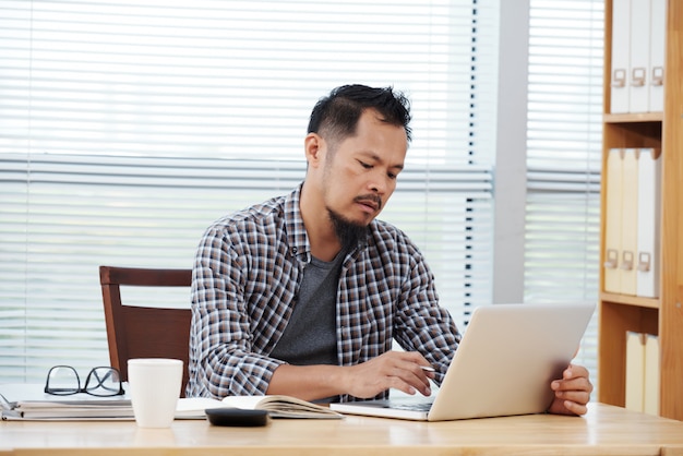 Casually dressed Filipino man sitting in office and working on laptop