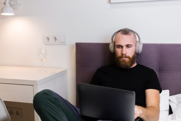 Caucasian bearded man in headphones in bedroom on bed working on laptop from home, typing, thinking.