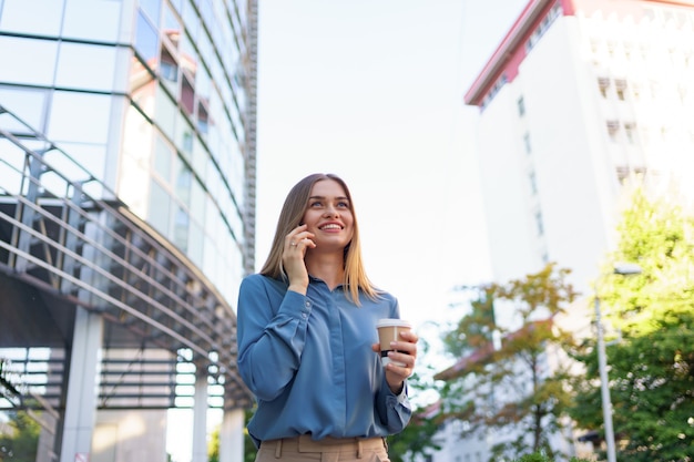 Caucasian business woman speaking by phone holding coffee to go. A successful European woman, talking on the phone, standing on modern office building