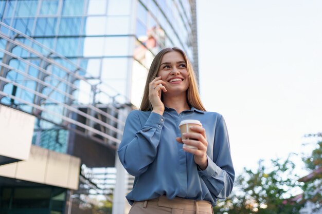 Caucasian business woman speaking by phone holding coffee to go. A successful European woman, talking on the phone, standing on modern office building