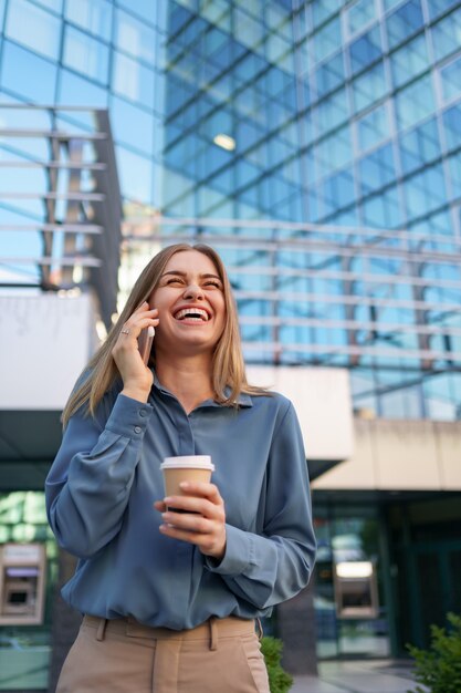 Caucasian business woman speaking by phone holding coffee to go. A successful European woman, talking on the phone, standing on modern office building