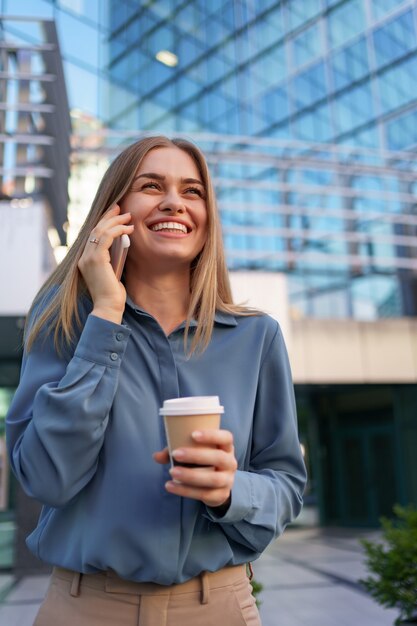 Caucasian business woman speaking by phone holding coffee to go. A successful European woman, talking on the phone, standing on modern office building