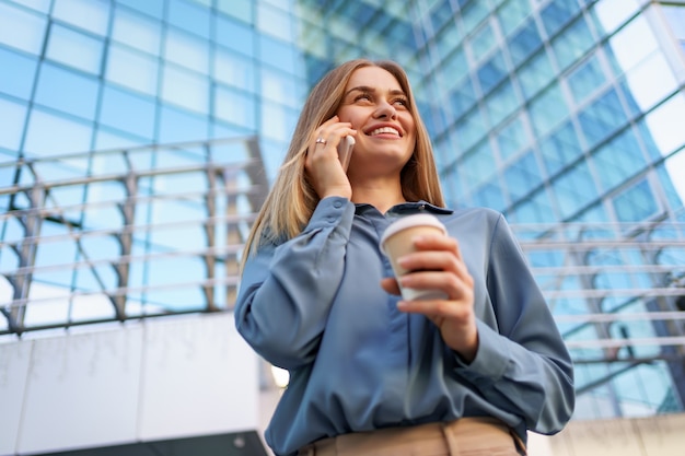 Caucasian business woman speaking by phone holding coffee to go. A successful European woman, talking on the phone, standing on modern office building