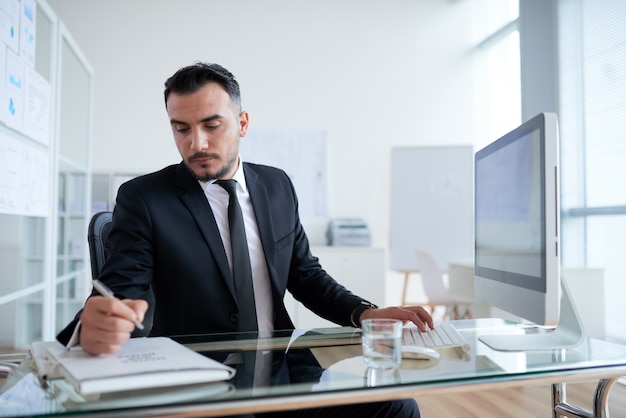 Caucasian businessman sitting at desk in front of computer and writing on document folder