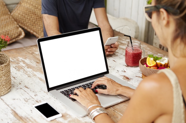 Free photo caucasian couple using free wi-fi at cafe and enjoying online communication on their electronic devices: woman checking email on laptop computer while her husband texting sms on mobile phone