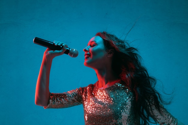 Caucasian female singer portrait isolated on blue studio background in neon light. Beautiful female model in bright dress with microphone. Concept of human emotions, facial expression, ad, music, art.