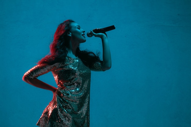 Caucasian female singer portrait isolated on blue studio background in neon light. Beautiful female model in bright dress with microphone. Concept of human emotions, facial expression, ad, music, art.